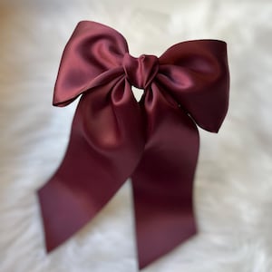 Peut inclure: Un grand nœud en satin bordeaux avec de longues queues flottantes. Le nœud est noué au centre et le tissu a un léger éclat. Le fond est une texture douce, blanche et duveteuse.