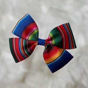 May include: A colorful hair bow featuring a striped pattern in shades of blue, red, green, and yellow. The bow is tied in the center with a blue ribbon, creating a vibrant accessory.