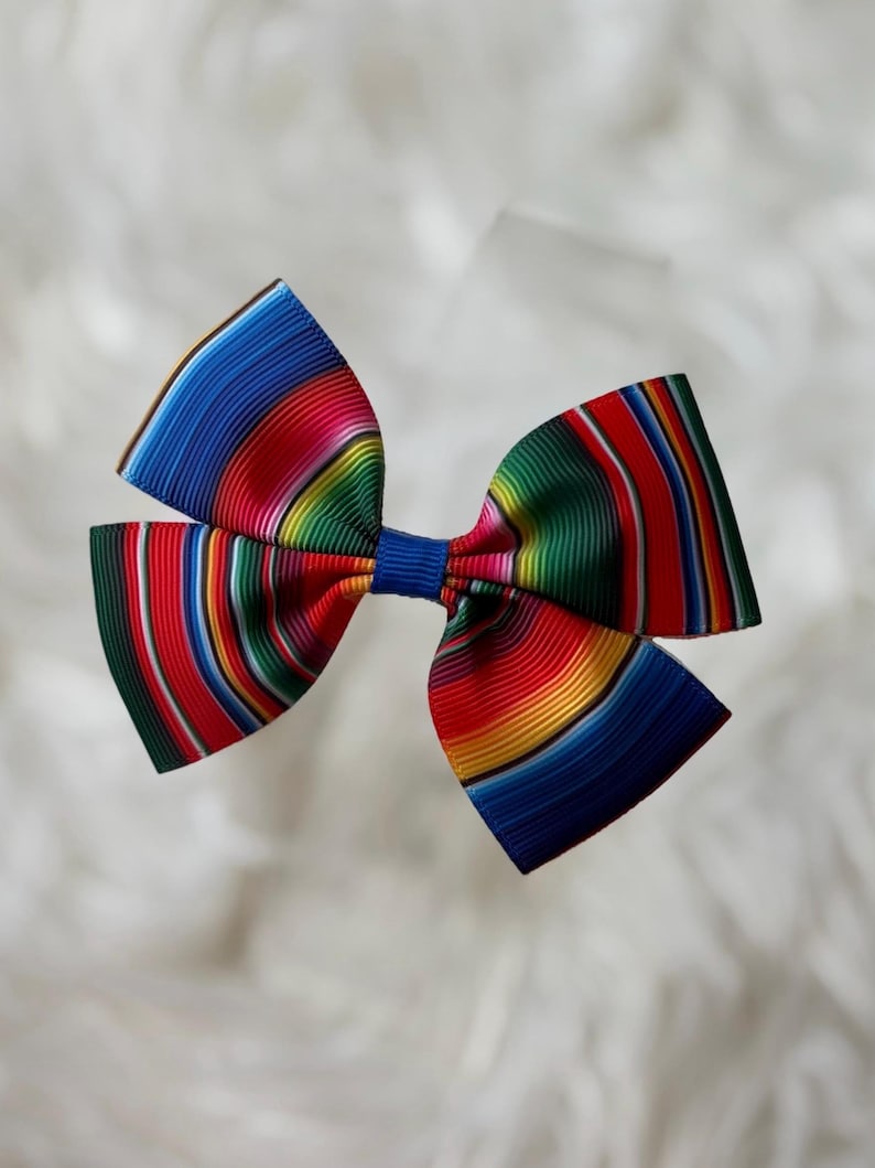 May include: A colorful hair bow featuring a striped pattern in shades of blue, red, green, and yellow. The bow is tied in the center with a blue ribbon, creating a vibrant accessory.