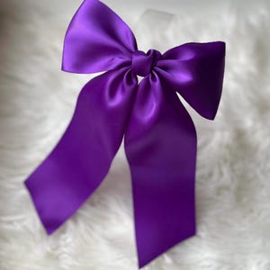 Könnte beinhalten: Eine leuchtend violette Satinschleife mit langen, fließenden Bändern. Die Schleife ist in der Mitte gebunden und bildet eine klassische Form. Der Stoff hat einen subtilen Glanz, der das Licht reflektiert. Diese dekorative Schleife eignet sich für verschiedene Bastelprojekte und Geschenkverpackungen.