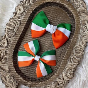 May include: Two hair bows with a green, white, and orange striped design, resembling a flag. The bows are on a decorative, ornate, brown tray with a white, fluffy background.