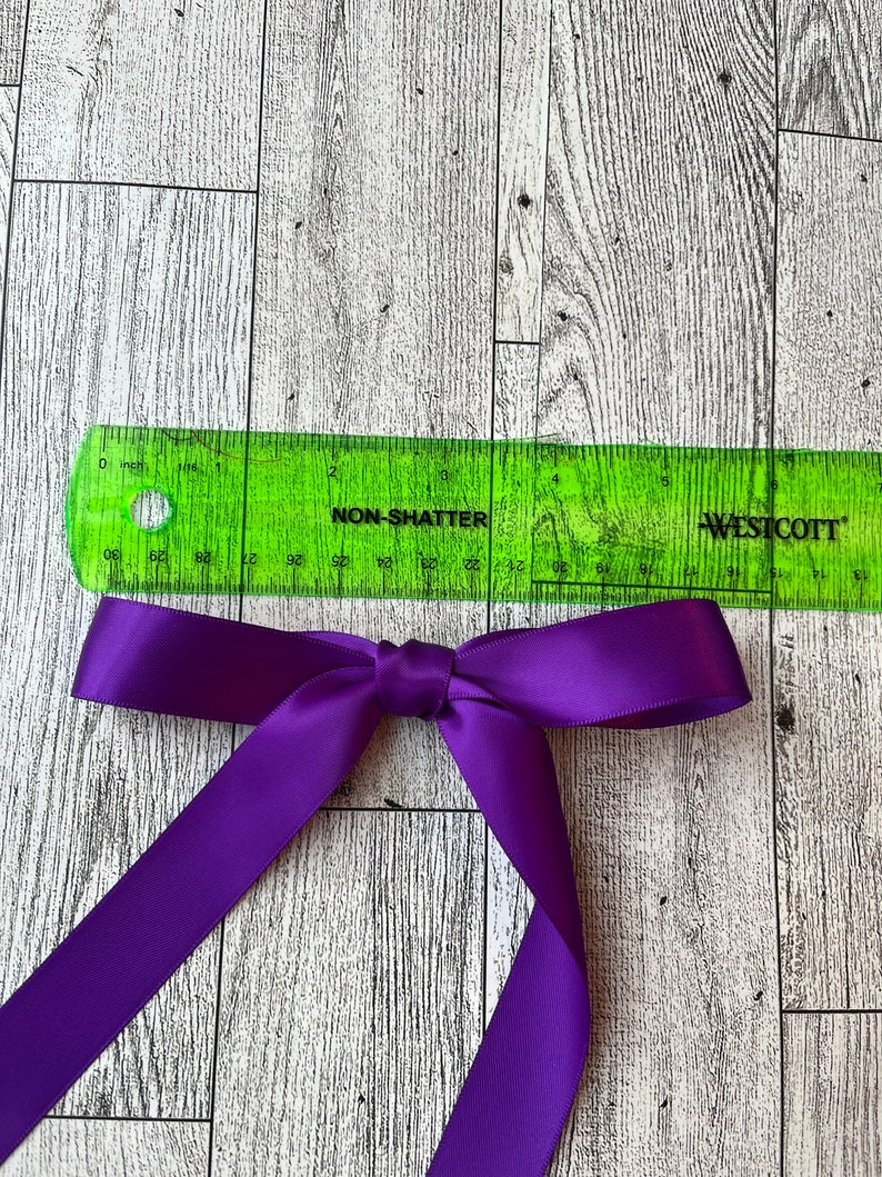 Może przedstawiać: Fioletowa wstążka satynowa zawiązana w kokardę, z zieloną linijką o długości 15 cm, na białym drewnianym tle.