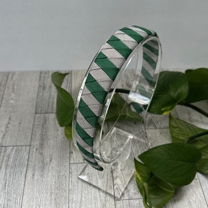 May include: A green and gray striped headband with a woven pattern. The headband is on a clear plastic stand.