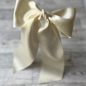 Könnte beinhalten: Eine große Schleife aus elfenbeinfarbenem Satin mit langen Bändern. Die Schleife ist zu einem Knoten gebunden und die Bänder fallen nach unten.