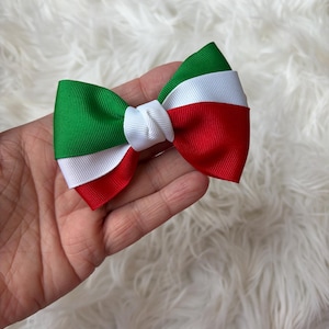 May include: A handmade bow tie featuring the colors of the Italian flag: green, white, and red. The bow is made of grosgrain ribbon and has a classic bow shape. The bow is held in a hand against a fluffy white background.