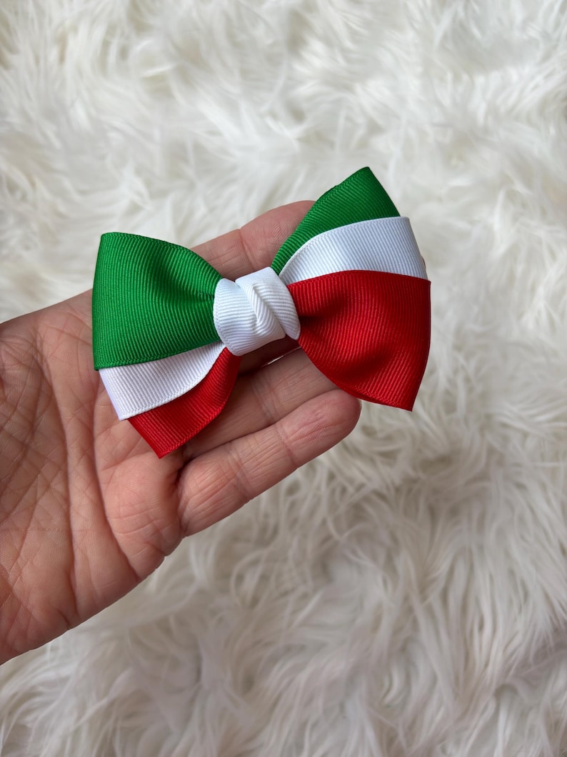 May include: A handmade bow tie featuring the colors of the Italian flag: green, white, and red. The bow is made of grosgrain ribbon and has a classic bow shape. The bow is held in a hand against a fluffy white background.