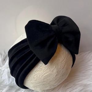 May include: A black velvet bow headband. The headband features a large, plush bow on top and a wide, ruched band. The bow and band are made of a soft, velvety material. The headband is displayed on a cream-colored, textured surface.
