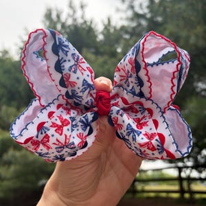 May include: A large, patriotic hair bow featuring a white background with red and blue bow and star patterns. The edges are trimmed with red and blue scalloped ribbon. The bow is held together with a red band.