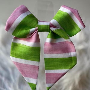 May include: A large, handmade bow with a striped pattern in pink, white, and green. The bow is tied in the center with long, flowing tails. The fabric appears to be a textured cotton or linen, perfect for hair accessories or gift wrapping.