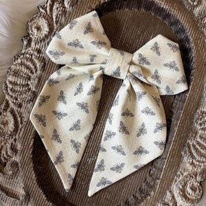 May include: A fabric hair bow featuring a cream-colored background with a repeating pattern of small, dark gray bees. The bow has two long, pointed tails and a central knot, constructed from a textured material with a visible weave pattern.