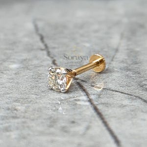 以下が含まれることがあります： 正方形の透明な宝石が付いた、金色のフラットバックスタッドピアス。 ピアスは4本爪セッティングです。 ピアスはグレーのテクスチャード加工された表面に置かれています。