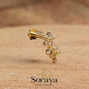 May include: A gold earring featuring a floral arrangement with small, clear gemstones. The earring is set against a textured, brown backdrop. The brand name "Soraya Fine Jewelry" is displayed below the earring.