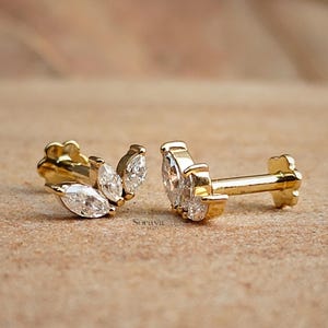 May include: Gold-colored stud earrings with a cluster of three marquise-cut, clear gemstones. The earrings have a floral-shaped backing and a post for piercing. The earrings are displayed on a light brown surface.