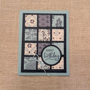Puede incluir: Una tarjeta de cumpleaños hecha a mano con un borde verde azulado y una cuadrícula de cuadrados estampados. Los cuadrados presentan diseños florales en tonos de azul, gris y crema. Un elemento circular en el centro dice "Happy Birthday" en escritura elegante.