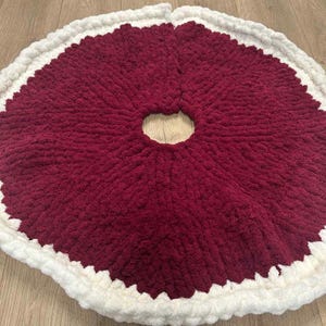 May include: A round, burgundy tree skirt with a white, scalloped edge. The skirt is made of a thick, plush material and has a circular opening in the center for the tree trunk. The overall design is simple and festive.
