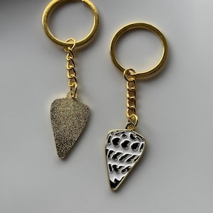 May include: Two gold keychains with teardrop-shaped pendants. One pendant has a textured, glittery gold surface. The other pendant features a black and white pattern with a gold border. Both keychains have gold chain links and gold key rings.
