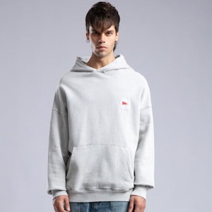 Op de afbeelding: Een persoon draagt een lichtgrijze hoodie met een voorzak en een kleine rood-witte patch. De persoon draagt ook lichtblauwe jeans en witte sneakers. De outfit is casual en modern.