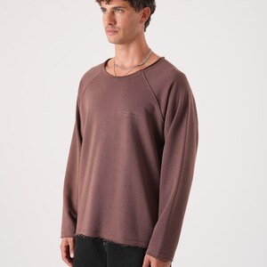 Può includere: Una maglietta marrone a maniche lunghe e girocollo con un piccolo logo discreto. La maglietta ha una vestibilità rilassata e dettagli con orlo grezzo. Il modello indossa jeans neri. Lo sfondo è bianco.