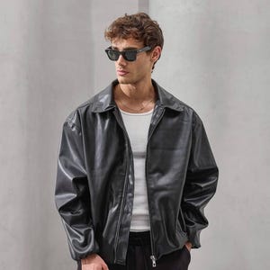 Op de afbeelding: Een zwarte imitatie leren jas met een kraag en een ritssluiting aan de voorkant. De jas wordt gedragen over een witte tanktop. De persoon draagt een zwarte zonnebril en een zwarte broek. De stijl is casual en modern.