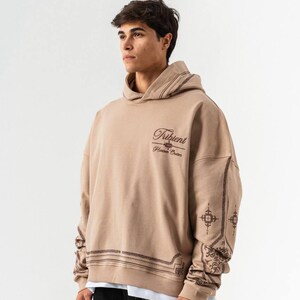 Puede incluir: Una sudadera con capucha de color beige con el texto "Tribient Hidden Codes" en la parte delantera. Las mangas y la capucha presentan intrincados diseños marrones. La sudadera se lleva sobre una camiseta blanca y con pantalones negros.