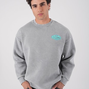 Pode incluir: Camisola cinzenta de gola redonda com aspeto desbotado. A camisola tem um pequeno patch oval no peito com o texto "FF Studio" em turquesa. O modelo está a usar calças de ganga escuras.