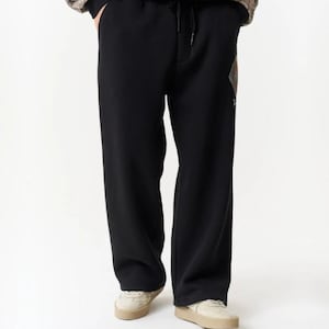 Può includere: Pantaloni della tuta neri con vita a coulisse e silhouette a gamba larga. I pantaloni sono abbinati a una felpa con cappuccio nera coordinata che presenta polsini decorati con un motivo stile paisley beige e nero.