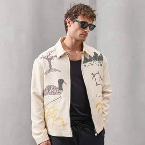 Op de afbeelding: Cr&egrave;mekleurige jas met illustraties met een natuurthema, waaronder een boom, een eend en een bergketen. De jas heeft een ritssluiting en wordt gedragen met een zwarte top en broek. Een uniek kledingstuk.
