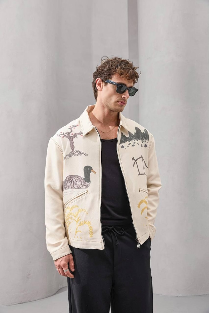 Op de afbeelding: Cr&egrave;mekleurige jas met illustraties met een natuurthema, waaronder een boom, een eend en een bergketen. De jas heeft een ritssluiting en wordt gedragen met een zwarte top en broek. Een uniek kledingstuk.