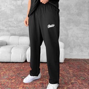Könnte beinhalten: Schwarze Jogginghose mit einem weißen "SWTP"-Logo auf dem linken Bein. Die Jogginghose hat einen Kordelzugbund und eine lockere Passform.