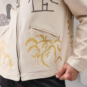 Op de afbeelding: Cr&egrave;mekleurige jas met ritssluiting met bruine en gouden illustraties van een eend, palmbomen en abstracte vormen. De jas heeft twee voorzakken en een zilveren ritssluiting.