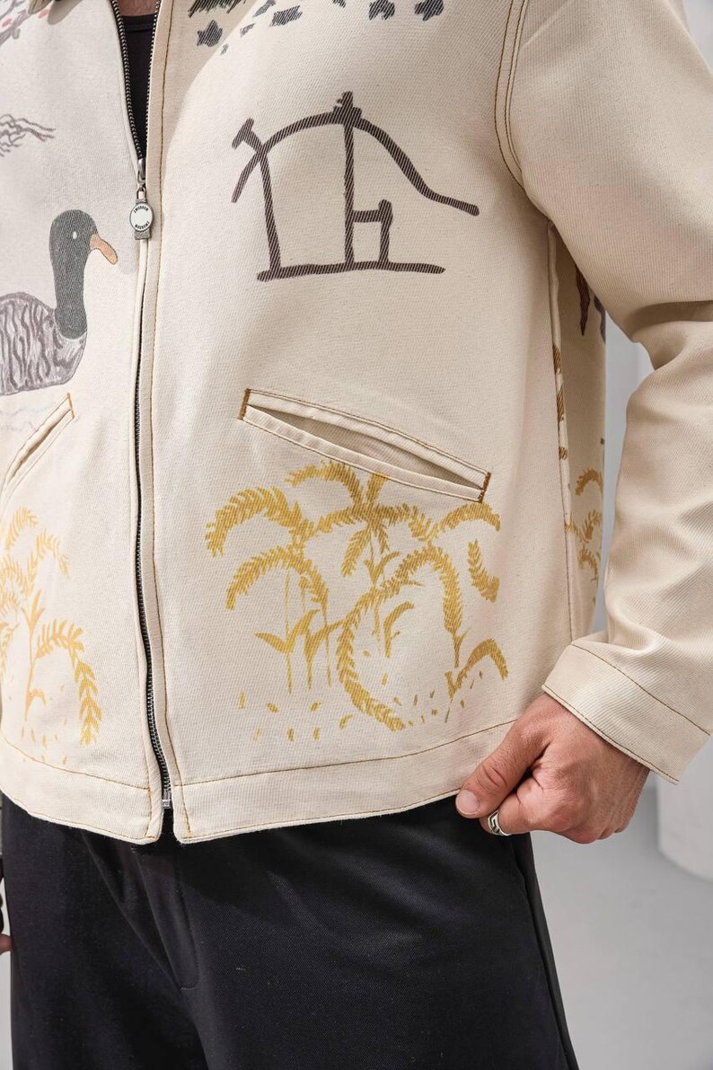 Op de afbeelding: Cr&egrave;mekleurige jas met ritssluiting met bruine en gouden illustraties van een eend, palmbomen en abstracte vormen. De jas heeft twee voorzakken en een zilveren ritssluiting.