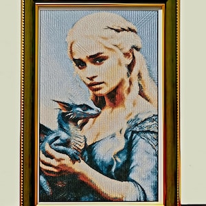 May include: Framed tapestry art featuring a woman with blonde hair holding a blue dragon. The artwork is in a gold and green frame. The woman is wearing a blue dress. The dragon is small and has blue scales.