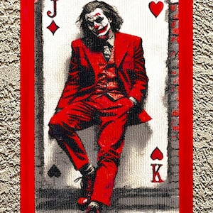 May include: Framed artwork depicting a figure in a red suit and white face paint, leaning against a playing card. The card shows a Jack of Diamonds, a Heart, a King of Hearts, and a Spade. The frame is red.