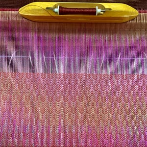 May include: A wooden loom with a partially woven textile. The textile features a gradient of pink and orange hues, with a patterned design. A wooden shuttle with a spool of thread is at the top. The loom is made of wood and metal.