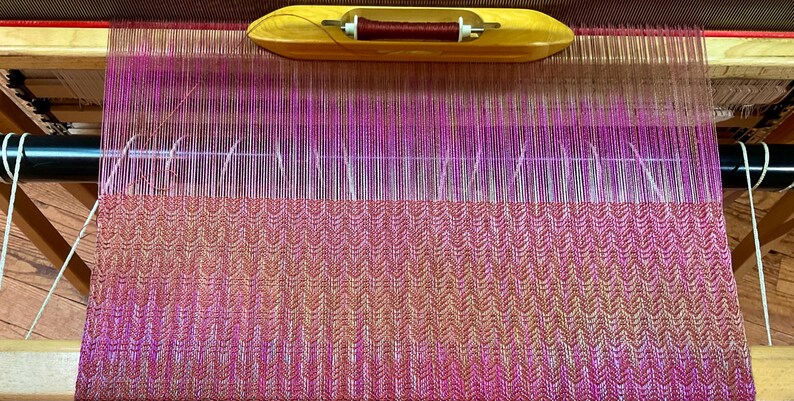 May include: A wooden loom with a partially woven textile. The textile features a gradient of pink and orange hues, with a patterned design. A wooden shuttle with a spool of thread is at the top. The loom is made of wood and metal.