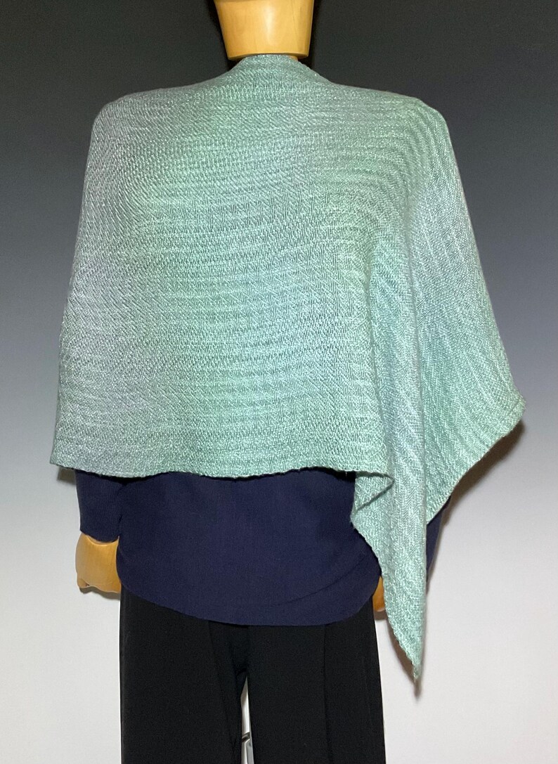 Pu&ograve; includere: Uno scialle tessuto verde chiaro con un motivo strutturato. Lo scialle &egrave; drappeggiato su un top blu navy e pantaloni neri. Lo scialle ha un aspetto morbido e fluido ed &egrave; esposto su un manichino.