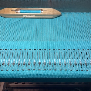 May include: A turquoise weaving project in progress on a wooden loom. A wooden shuttle holds a spool of turquoise thread. The woven fabric features a decorative border with a repeating pattern of white and brown elements.