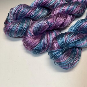 May include: Close-up of several skeins of yarn in shades of purple, blue, and silver. The yarn is tightly wound, showing the texture and color variations. The yarn is likely for knitting or crocheting projects.