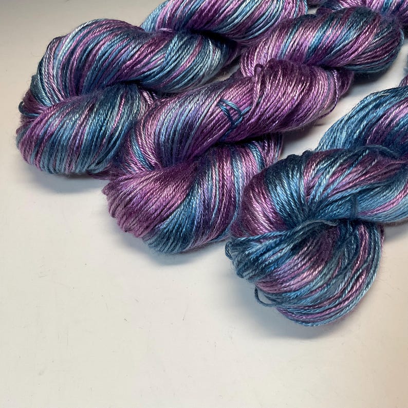 May include: Close-up of several skeins of yarn in shades of purple, blue, and silver. The yarn is tightly wound, showing the texture and color variations. The yarn is likely for knitting or crocheting projects.