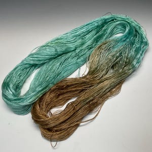 Puede incluir: Dos madejas de hilo en la imagen. Una es de color verde azulado claro, y la otra es una mezcla de oro y verde oliva. El hilo parece ser una fibra fina y sedosa, adecuada para proyectos delicados como tejer o hacer ganchillo.
