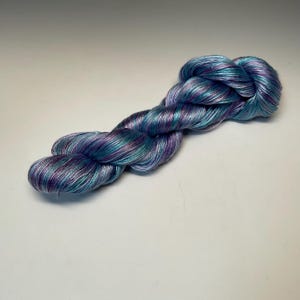 May include: A skein of hand-dyed yarn in shades of blue, teal, and purple. The yarn is twisted into a braid shape, showcasing the color variations. The yarn appears soft and has a slight sheen, suitable for knitting or crochet projects.