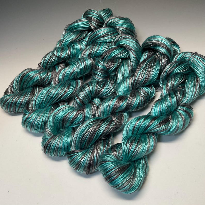 Op de afbeelding: Verschillende strengen garen in tinten van teal, grijs en zilver zijn gerangschikt op een wit oppervlak. Het garen is strak getwist, waardoor een textuur ontstaat. De kleurencombinatie geeft het garen een glinsterend effect, geschikt voor brei- of haakprojecten.