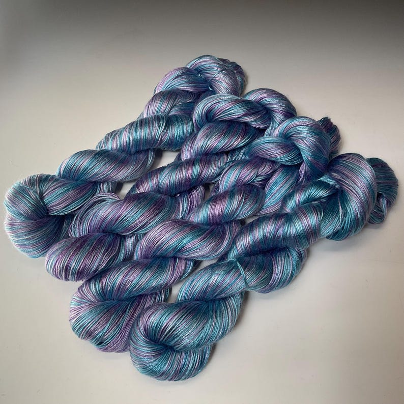 May include: Several skeins of hand-dyed yarn in shades of blue, teal, and purple. The yarn is twisted into neat bundles, showcasing the color variations. The yarn has a subtle sheen, suggesting a soft, luxurious fiber.