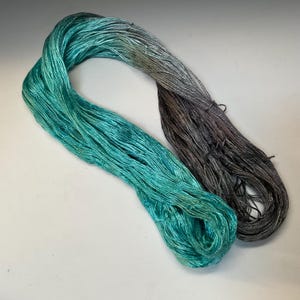 Op de afbeelding: Een streng garen in tinten van turquoise en grijs. Het garen is fijn gesponnen en heeft een lichte glans, wat wijst op zijde of een vergelijkbare vezel. De kleuren gaan soepel over van een levendig turquoise naar een gedempt grijs, waardoor een gradi&euml;nteffect ontstaat.