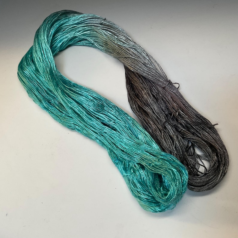 Op de afbeelding: Een streng garen in tinten van turquoise en grijs. Het garen is fijn gesponnen en heeft een lichte glans, wat wijst op zijde of een vergelijkbare vezel. De kleuren gaan soepel over van een levendig turquoise naar een gedempt grijs, waardoor een gradi&euml;nteffect ontstaat.