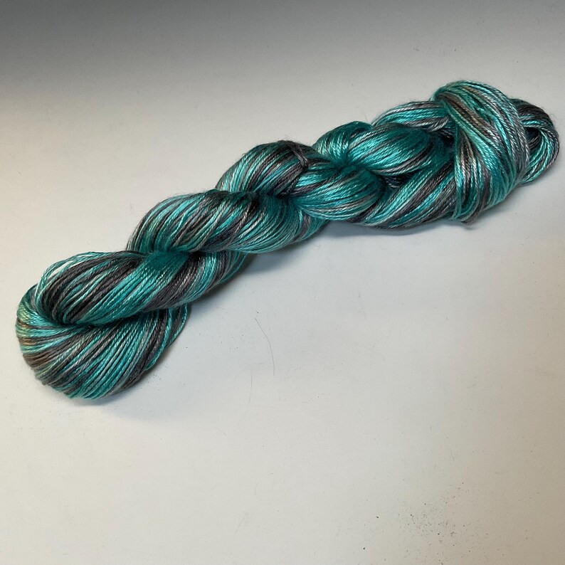 Op de afbeelding: Een streng handgeverfd garen in tinten van turquoise, grijs en wit. Het garen is tot een vlecht gedraaid, waarbij de kleurvariaties worden getoond. Dit garen is geschikt voor brei- en haakprojecten.