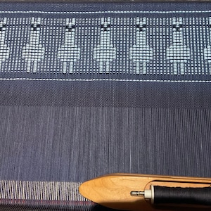 May include: A close-up of a loom with a blue and white woven pattern. A wooden shuttle with a spool of black thread is in the foreground.