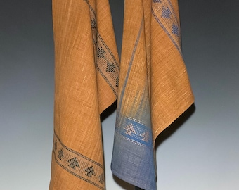 May include: Two brown linen towels with decorative blue and black geometric patterns hanging from a wooden rod. One towel has a blue gradient. The towels are made of a textured fabric.