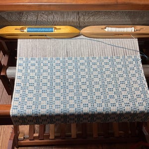 Puede incluir: Un telar de madera con una tela tejida que presenta un patrón de cuadros azules y blancos. Dos lanzaderas de madera descansan sobre los hilos de urdimbre, una con una bobina de hilo azul y la otra con una bobina de hilo blanco.