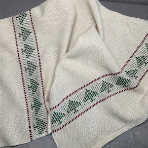 May include: A white linen tea towel with green and red woven borders. The borders feature a repeating pattern of small green leaves.